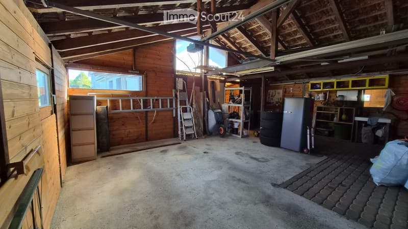 Empty garage with wooden walls, concrete floor, and exposed wooden beams on the ceiling. Contains storage shelves, ladders, and tools, but no furniture.