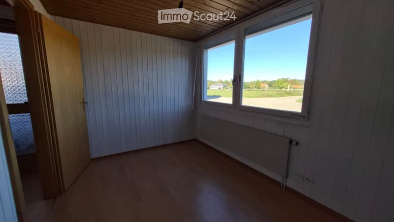 Empty room with wooden flooring, white paneled walls, large windows offering a view of the countryside, and a radiator under the windows.