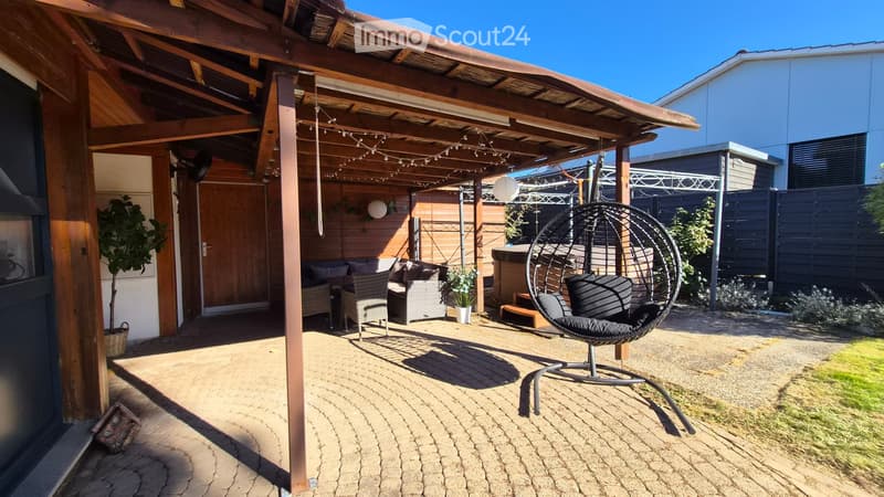 A covered outdoor patio with a sofa, chairs, a wicker hanging chair, a small table with a flower pot, and a wooden fence.