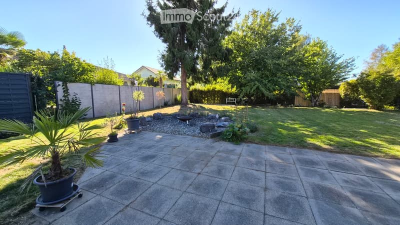 Well-maintained garden with a paved patio, potted plants, a small seating area, and a grassy lawn surrounded by trees and a wooden fence.
