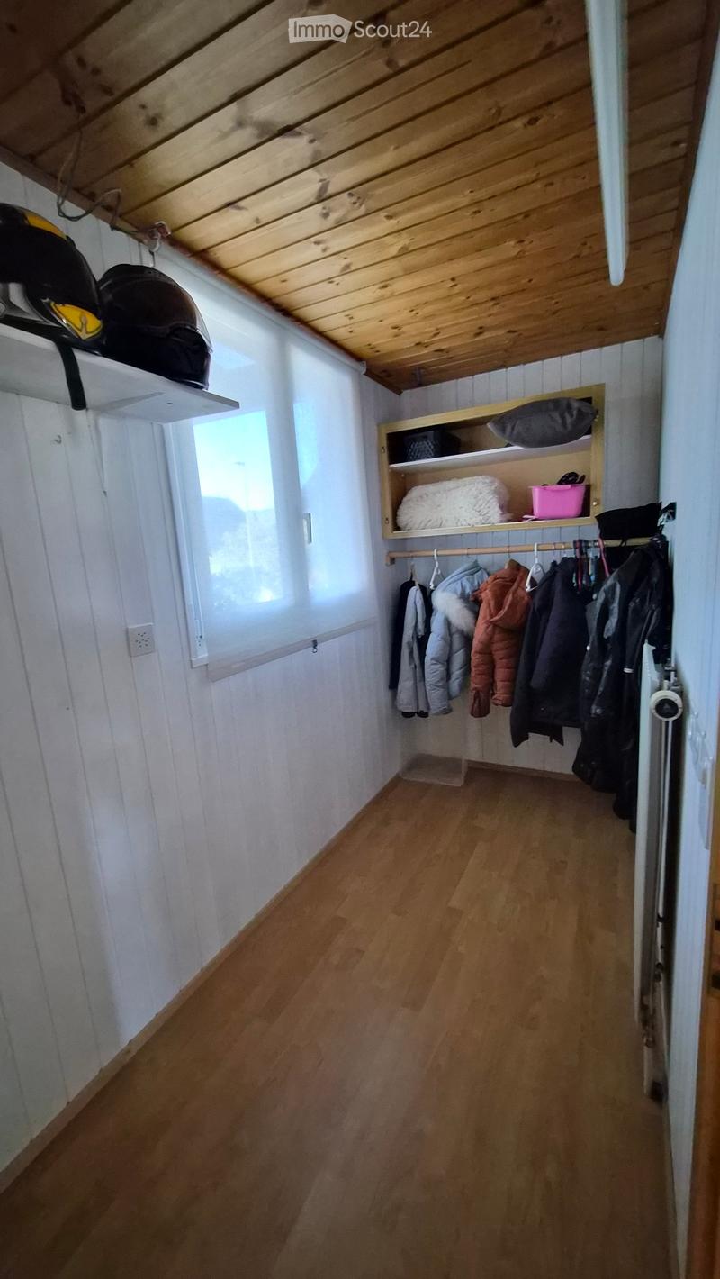A narrow hallway with wooden flooring, white walls, and a slanted wooden ceiling. Features include built-in shelves with folded blankets, a hanging rack with jackets, and a small window allowing natural light.