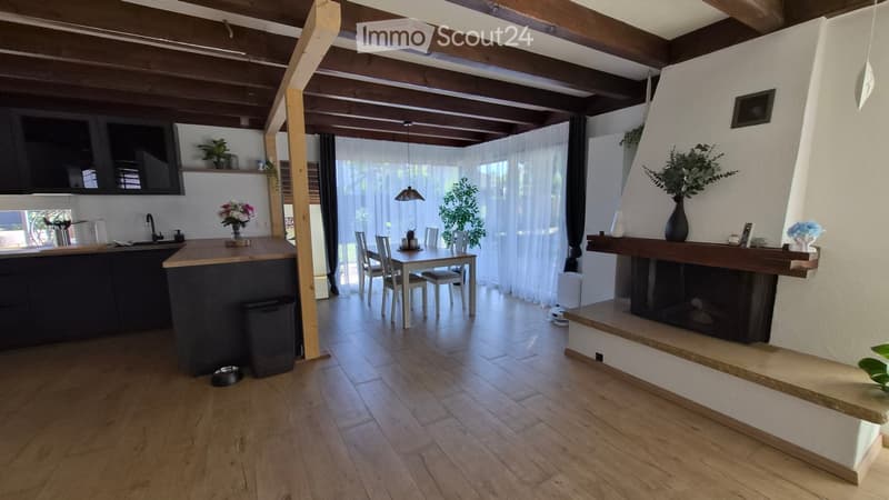 Open-plan living and dining area with wooden ceiling beams, a kitchen island, a dining table with chairs, a fireplace, and a TV screen.