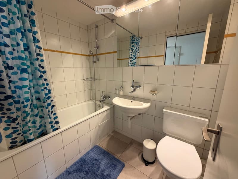 Modern bathroom with white tiles, bathtub, sink, toilet, mirror, and a blue bath mat.