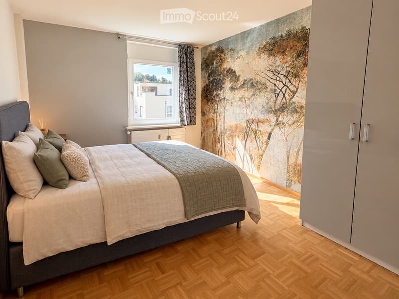 Modern bedroom with a large bed, multiple pillows, a grey blanket, a large abstract wall mural, and a window with a curtain. The room features wooden flooring and a built-in wardrobe.