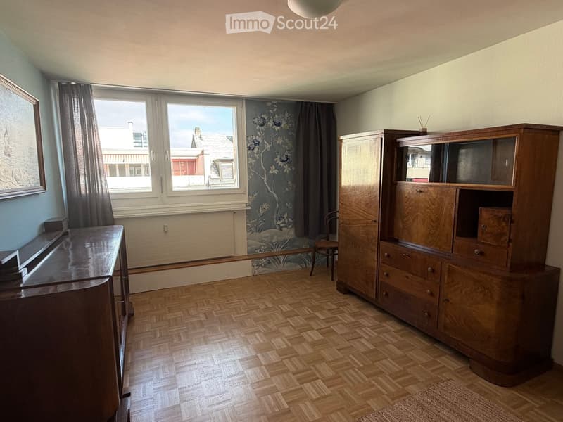 Empty room with wooden herringbone flooring, large window with floral wallpaper, wooden cabinet, and a small chair near the window.