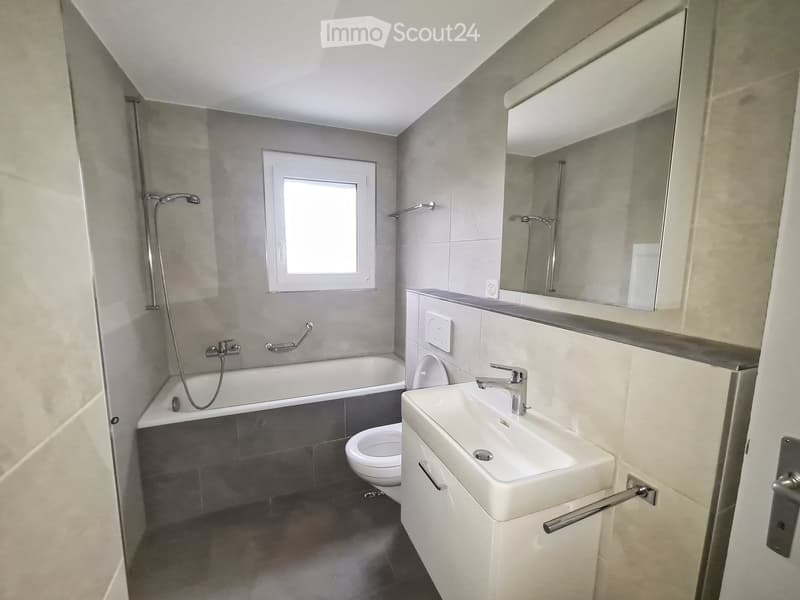 Modern bathroom with a bathtub, toilet, sink, mirror, and showerhead. Features a window and sleek, contemporary design.