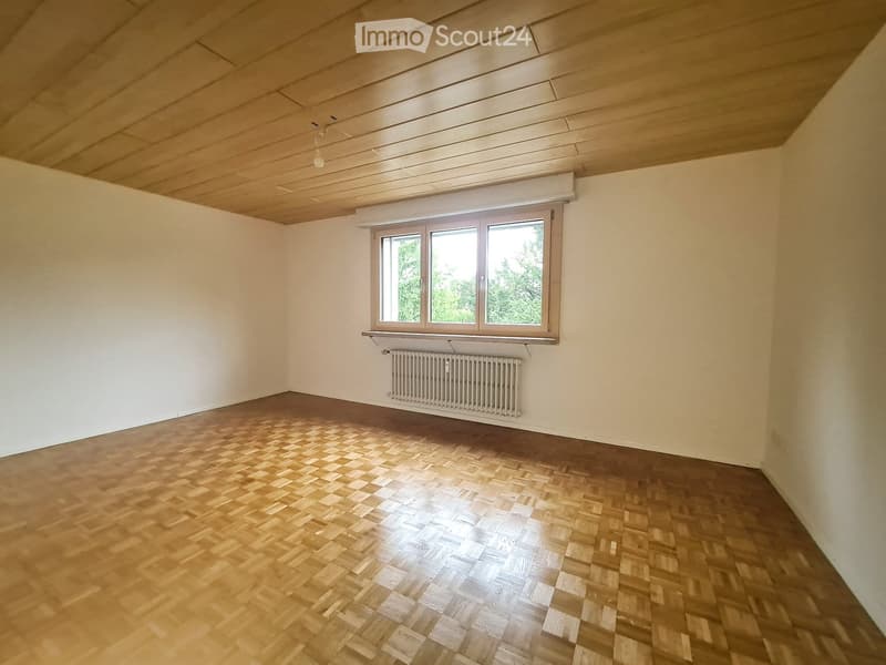 Empty room with wooden herringbone flooring, white walls, and a large window with a radiator below it.