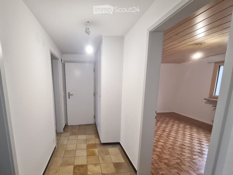 Empty hallway with white walls, tiled floor, and wooden flooring in the adjacent room. Features a hanging light fixture and several doors.