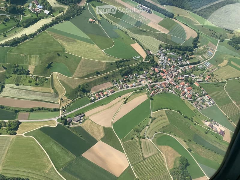 Aerial view of a rural area with scattered houses, fields, and roads. The landscape includes green fields, patches of forest, and a small village with red-roofed buildings.