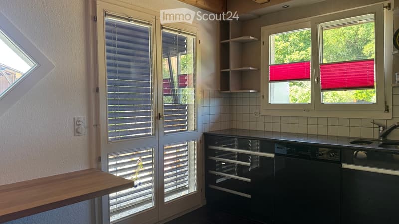 Modern kitchen with white tiled walls, dark countertops, built-in appliances including a dishwasher and oven, and a wooden table near the window.