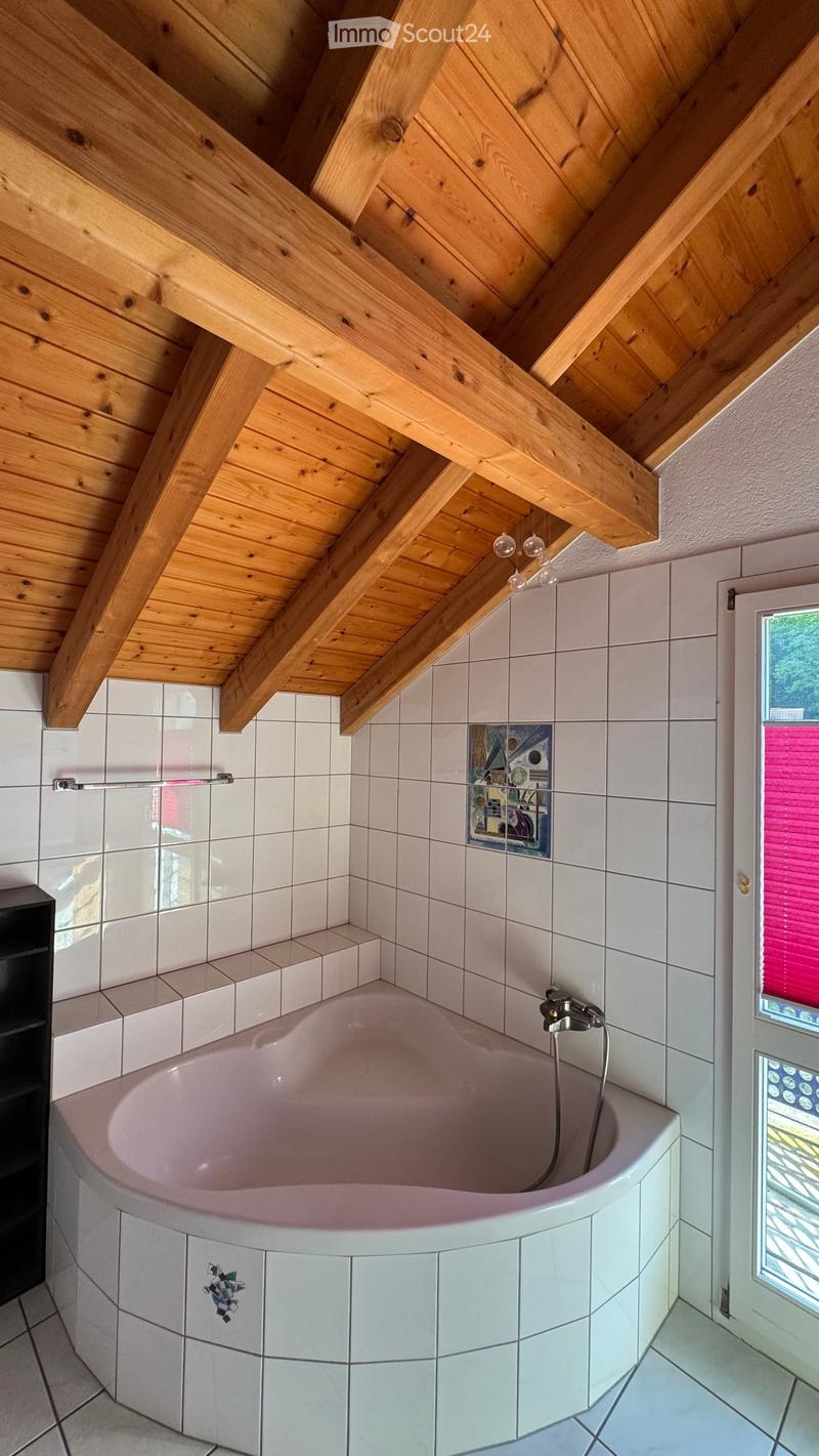 Bathroom with a wooden slanted ceiling, a bathtub, a mirror, and a shelf. The walls are tiled, and there is a window with blinds.