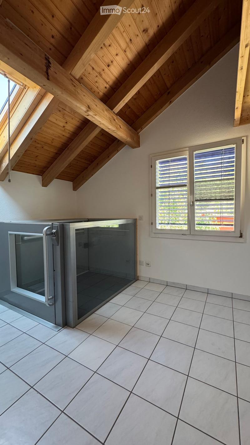 Empty room with white tiled flooring, exposed wooden beams on the ceiling, and a glass-enclosed elevator shaft. Two windows with blinds are visible on the right side.