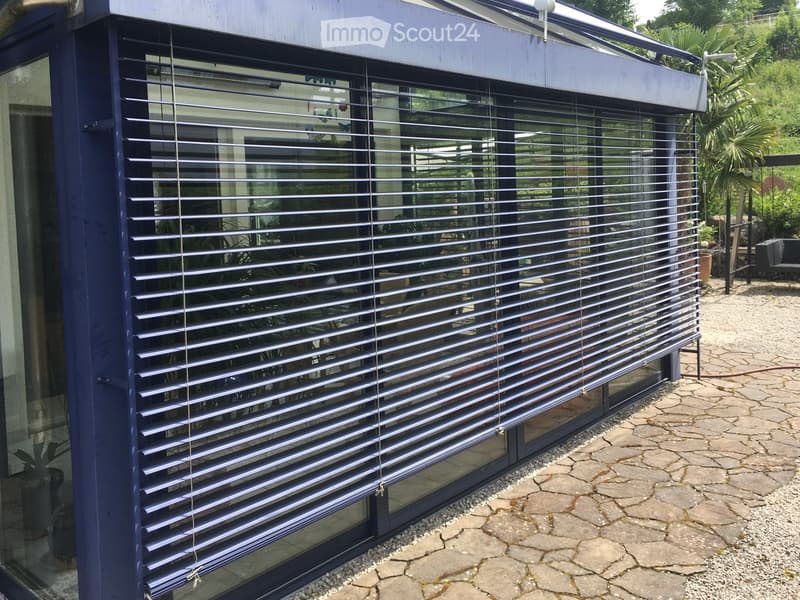 A modern balcony or terrace with blue roller blinds, stone pavement, and surrounding greenery.