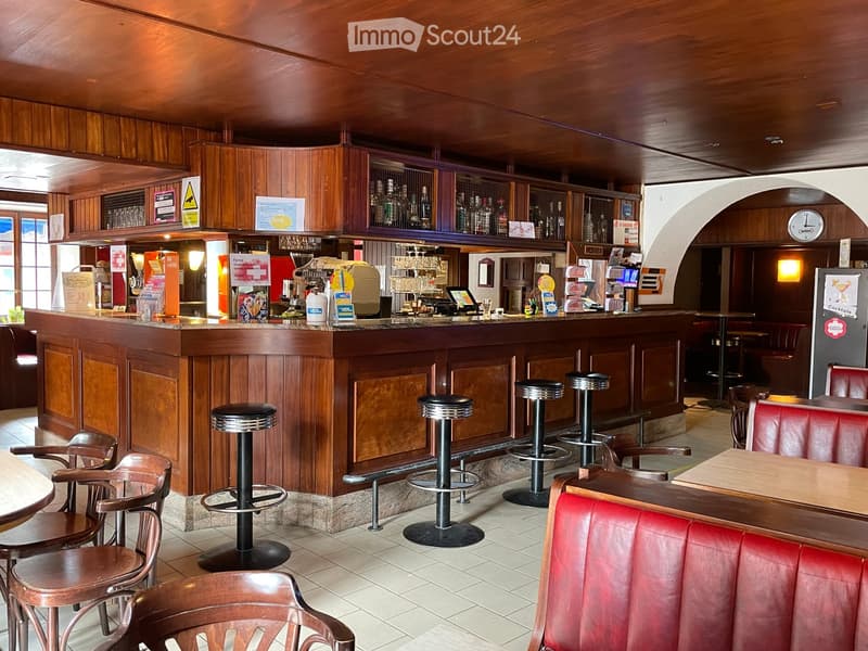 A cozy bar area with wooden paneling, a central bar counter, several high stools, tables, and chairs, along with shelves stocked with bottles and glasses.