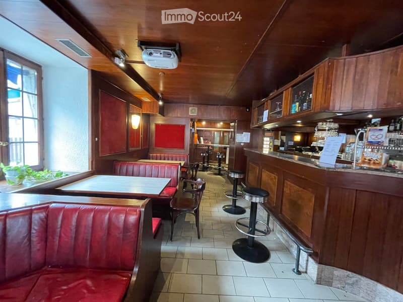 Restaurant interior with red leather booths, wooden tables, chairs, and a bar counter with stools. Wooden ceiling and walls, large windows, and a projector mounted on the ceiling.