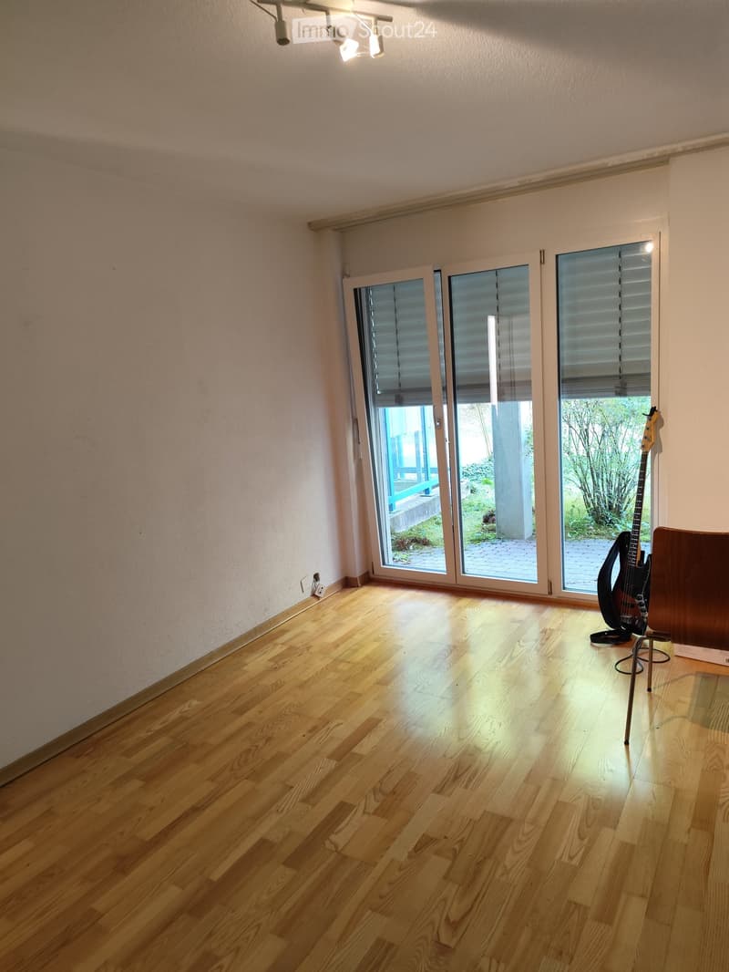 Empty room with wooden flooring, large sliding glass doors leading to an outdoor area, and a single guitar leaning against the wall.