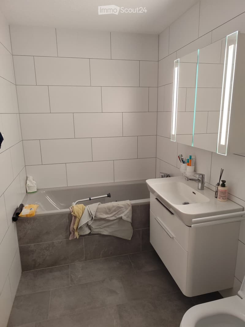 Modern bathroom with white tiles, a sink with a mirror, a bathtub, and a toilet. Towels are draped over the edge of the bathtub.
