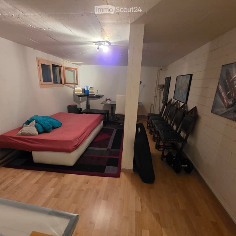 A small, furnished room with a bed, table, chair, and several picture frames on the wall. The room has wooden flooring and a window with blinds.