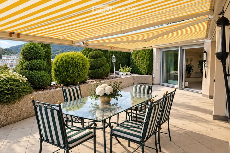 A covered outdoor terrace with a glass dining table, four striped chairs, and a vase of white flowers. The terrace has a yellow and white striped awning, tiled flooring, and overlooks a landscaped garden with trimmed bushes and trees.