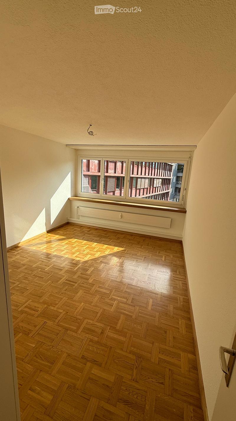 Empty room with wooden herringbone flooring, white walls, and a large window with a radiator below it.