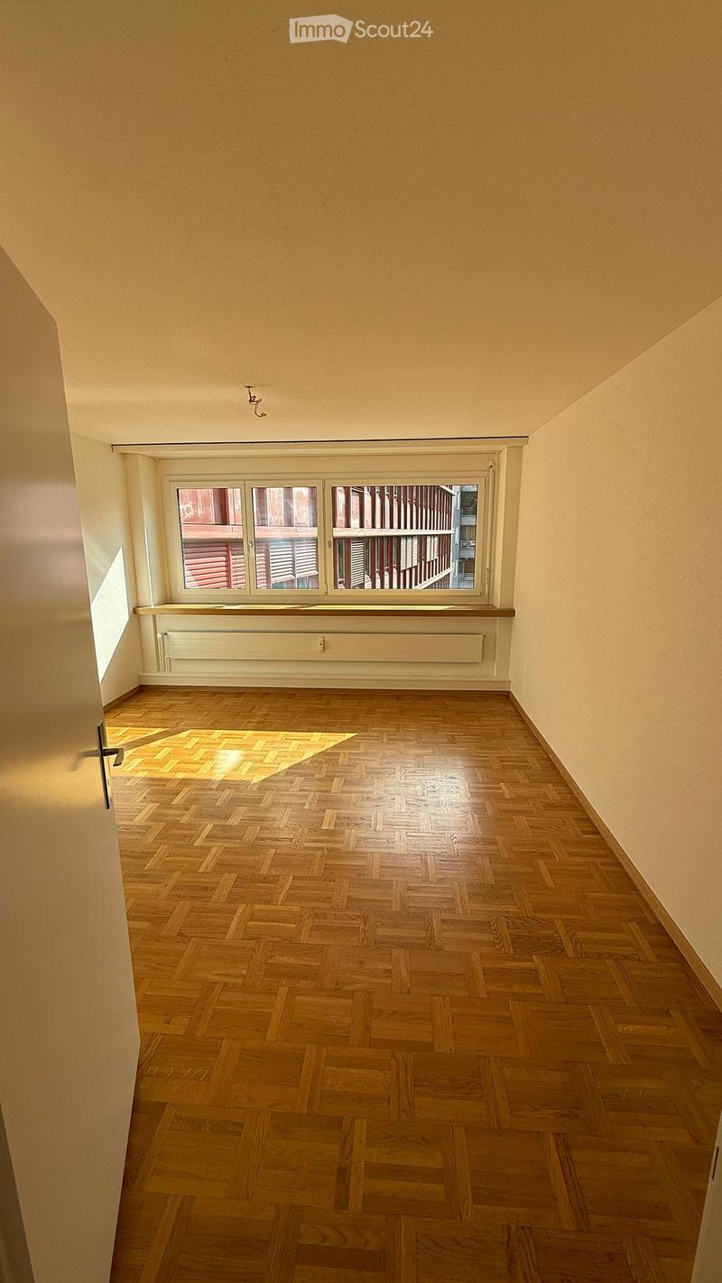 Empty room with wooden herringbone flooring, light-colored walls, and a large window with blinds. No furniture or equipment is present.