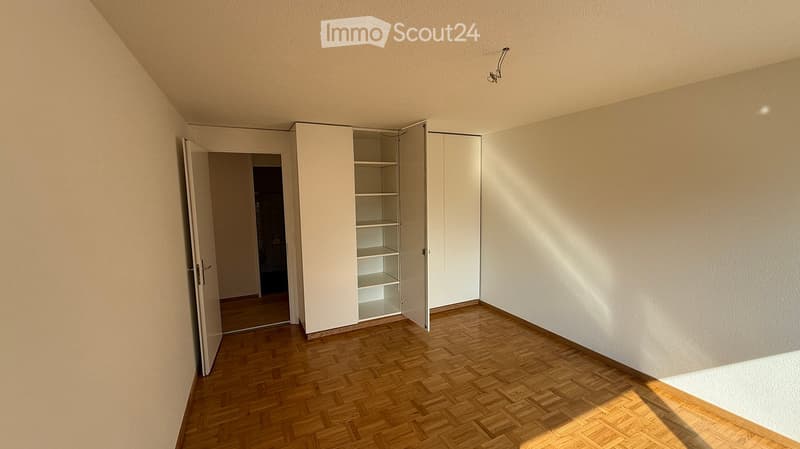 Empty room with wooden flooring, white walls, and a built-in closet with empty shelves. Sunlight streams in from the right side.