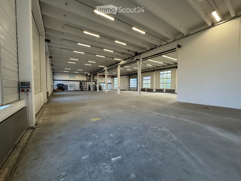 Large, empty industrial-style room with concrete floors, white walls, exposed ceiling beams, and large windows allowing natural light.