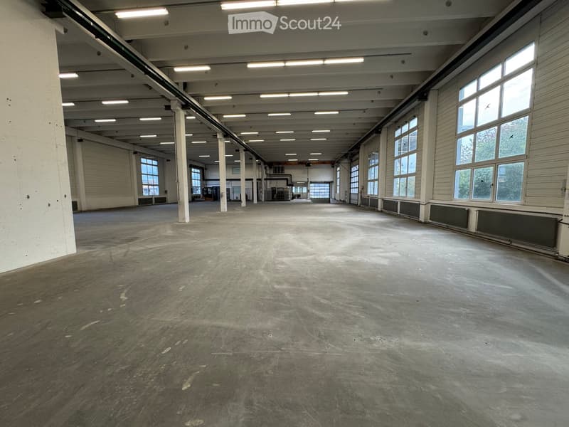 Large, empty industrial-style room with concrete floors, exposed metal beams, and large windows allowing natural light. No furniture or equipment is present.