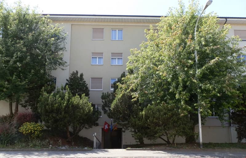 A multi-story residential building with beige walls, multiple windows, and a small entrance area surrounded by trees and greenery.