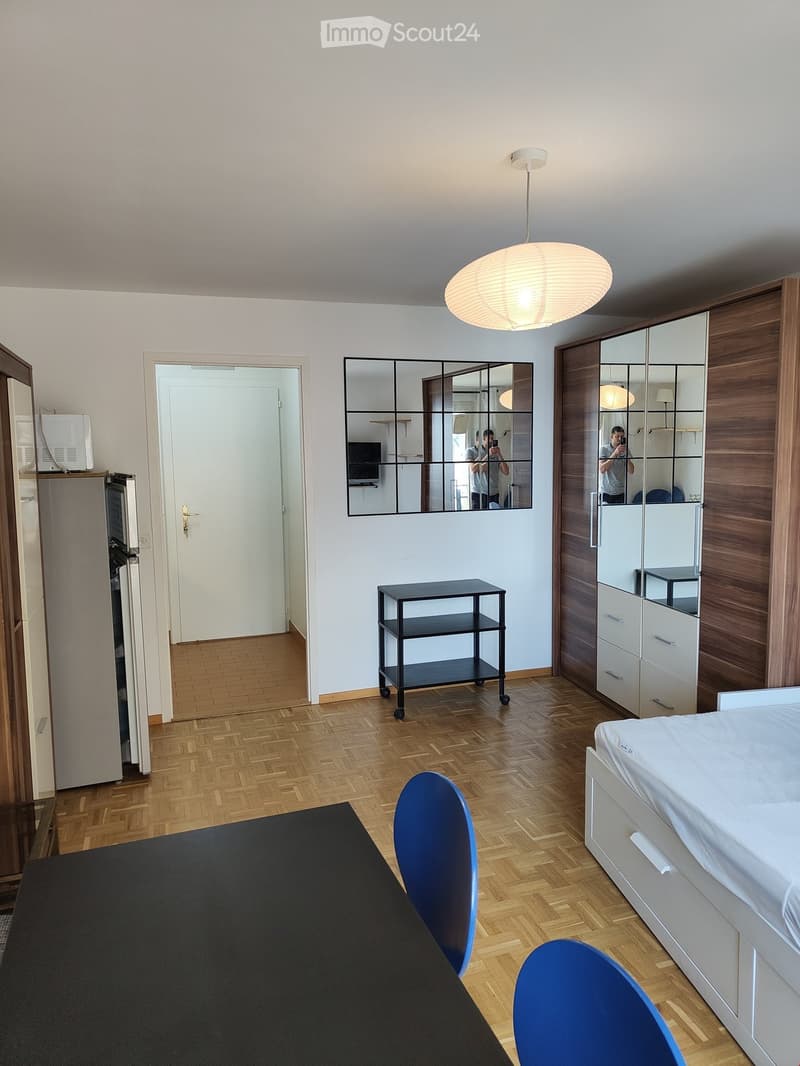A modern, well-lit room with a bed, a black rolling table, a large mirror, a refrigerator, and a wooden cabinet. The floor is made of herringbone-patterned wood, and the walls are painted white.