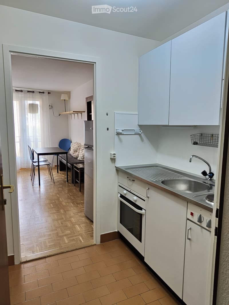 The image shows a modern kitchen with white cabinets, a stainless steel sink, and an oven. A small dining area is visible through an open doorway, featuring a table, chairs, and a bench with cushions.