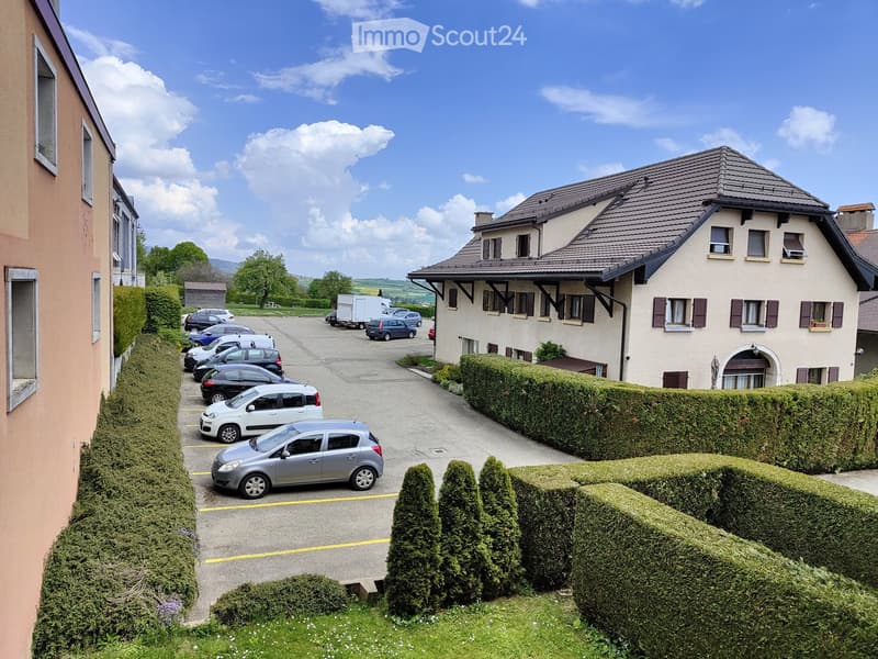 Two-story residential building with a tiled roof, multiple windows, and a well-maintained hedge-lined driveway with several parked cars.
