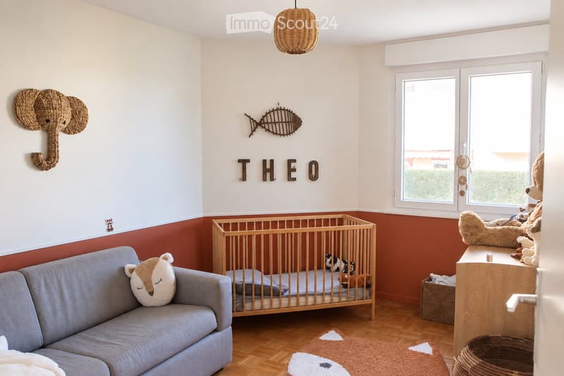 A cozy children's bedroom with a grey sofa, wooden crib, plush toys, a wicker basket, and a wicker pendant light. The room features a warm color palette with a red accent wall and a large window offering natural light.