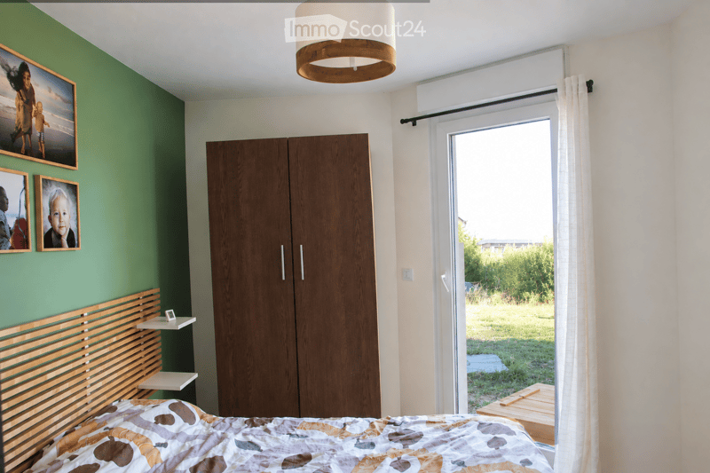 Modern bedroom with a wooden bed frame, a large wardrobe, and a window with white curtains. The room features a colorful bedspread and several picture frames on the wall.