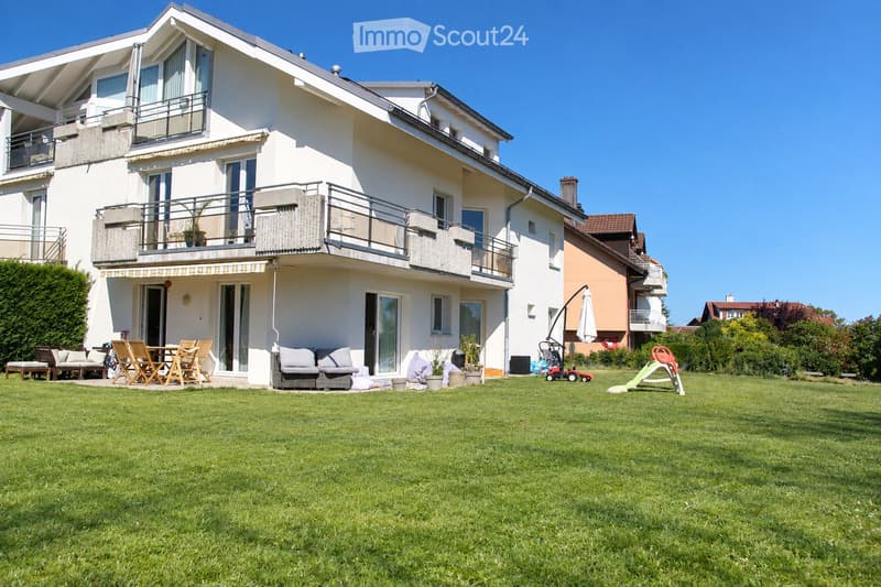 Two-story house with white exterior, multiple balconies, large windows, and a well-maintained lawn with outdoor furniture and play equipment.