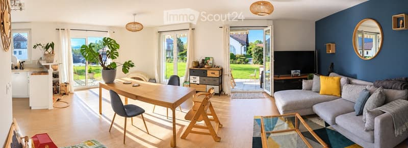 Bright living room with grey sectional sofa, wooden coffee table, dining table with chairs, large mirror, and potted plant. The room features a TV stand and a storage unit, with large windows and sliding glass doors leading to a patio.