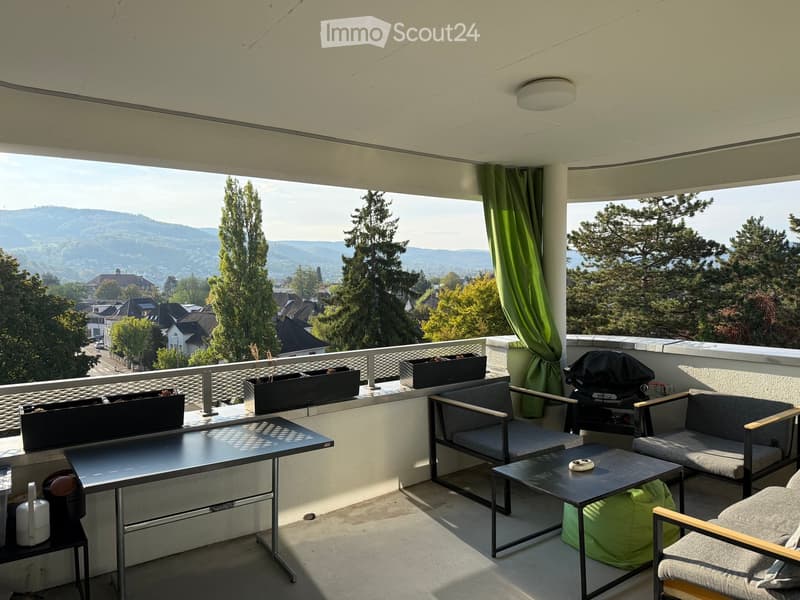 Balcony with a scenic view of a town and mountains, featuring a sofa, chairs, a table, a grill, and flower pots.