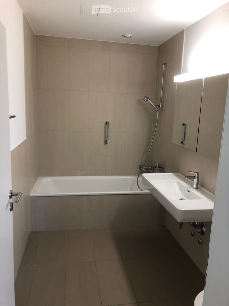 Modern bathroom with beige tiled walls and floor, featuring a bathtub, a sink with a modern faucet, a mirror, and a showerhead with a handheld hose.