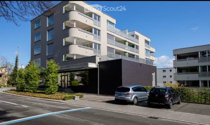 Modern multi-story residential building with balconies and a covered entrance, two parked cars in front.