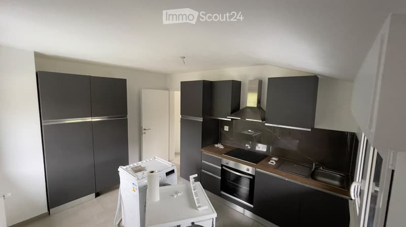 Modern kitchen with dark grey cabinets, stainless steel appliances, and a white washing machine. The space includes a sink, oven, and refrigerator, with a small table holding a paper towel roll.