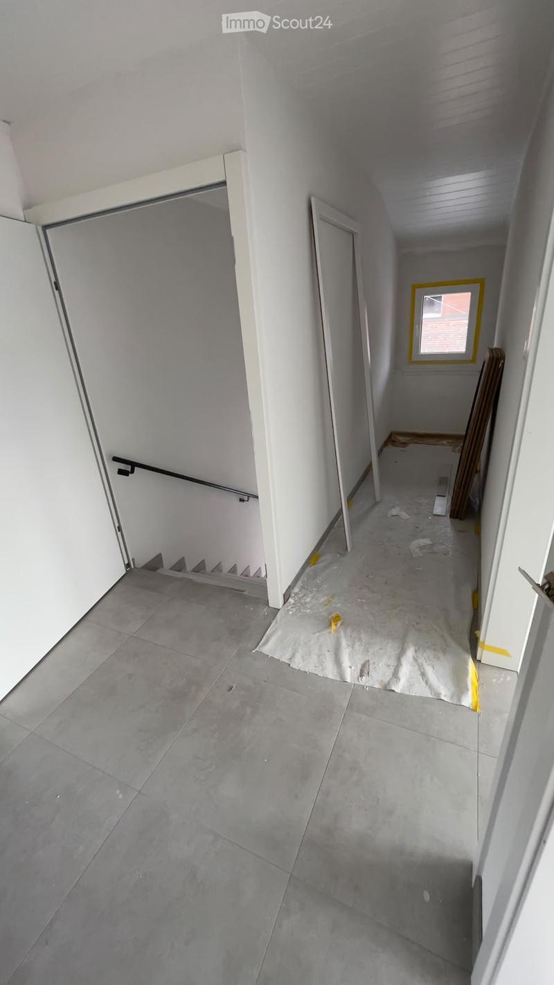 Empty hallway with grey tiled floor, white walls, and a staircase leading upwards. A small window with yellow frame is visible at the end of the hallway.