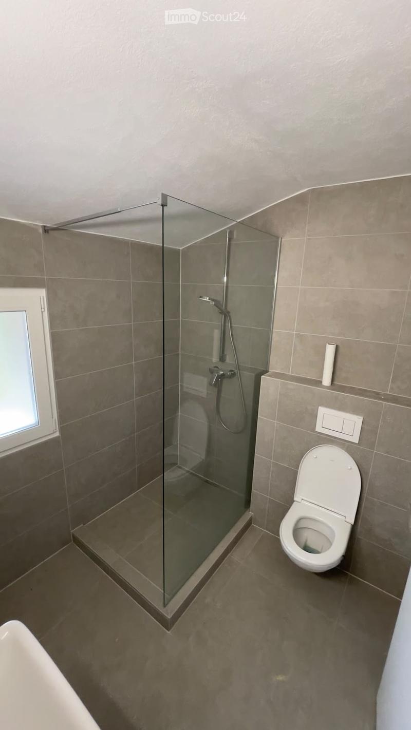Modern bathroom with a glass-enclosed shower, toilet, and a small window. The walls and floor are tiled in a neutral gray color.