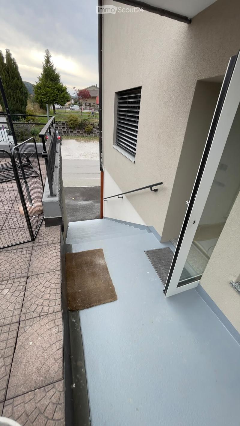 Balcony with light blue flooring, a brown doormat, a metal railing, and a window with blinds. The balcony is adjacent to a building with a beige exterior wall.