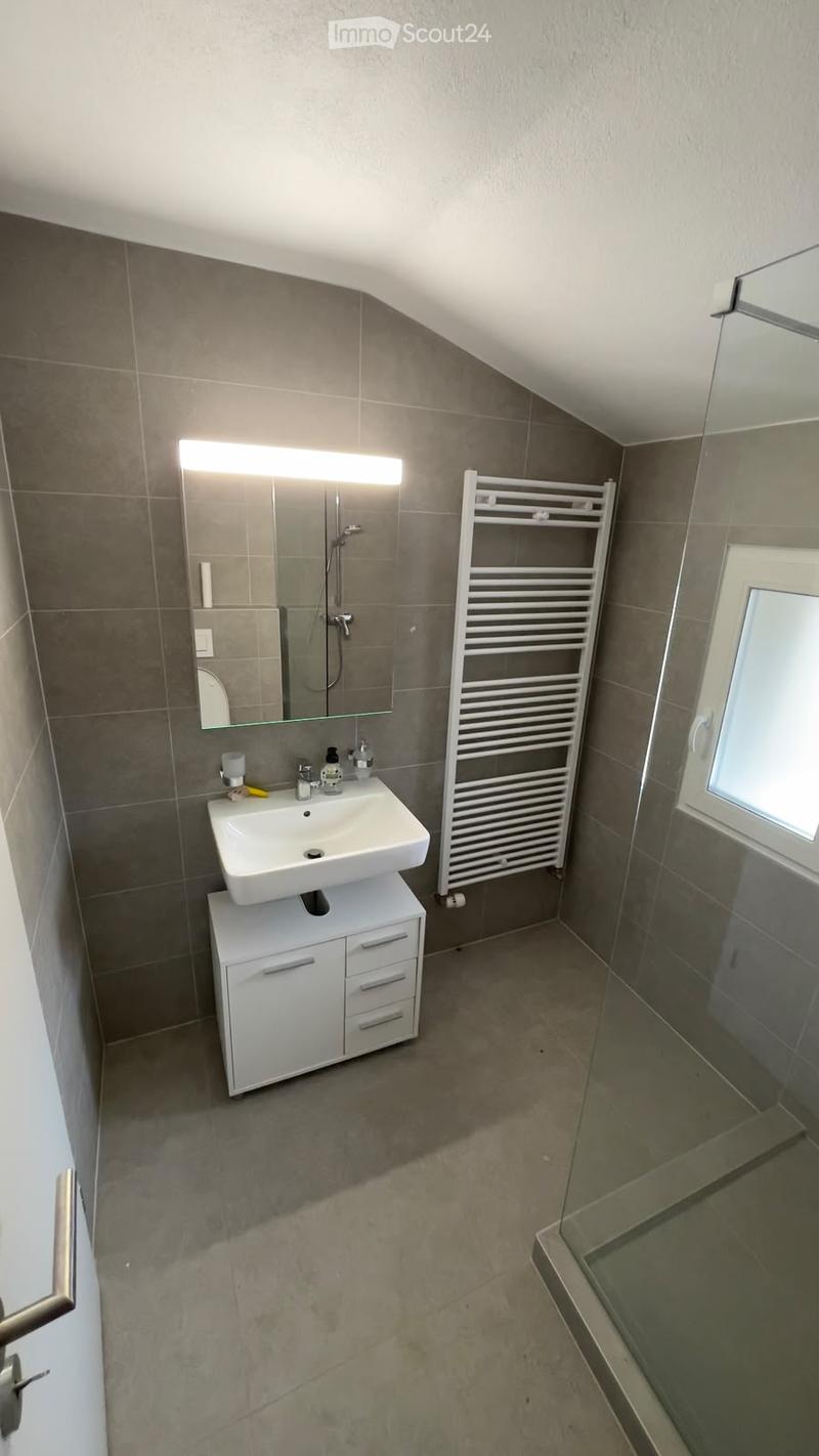 Modern bathroom with grey tiles, white sink, mirror with built-in lighting, towel rack, and a glass-enclosed shower area.