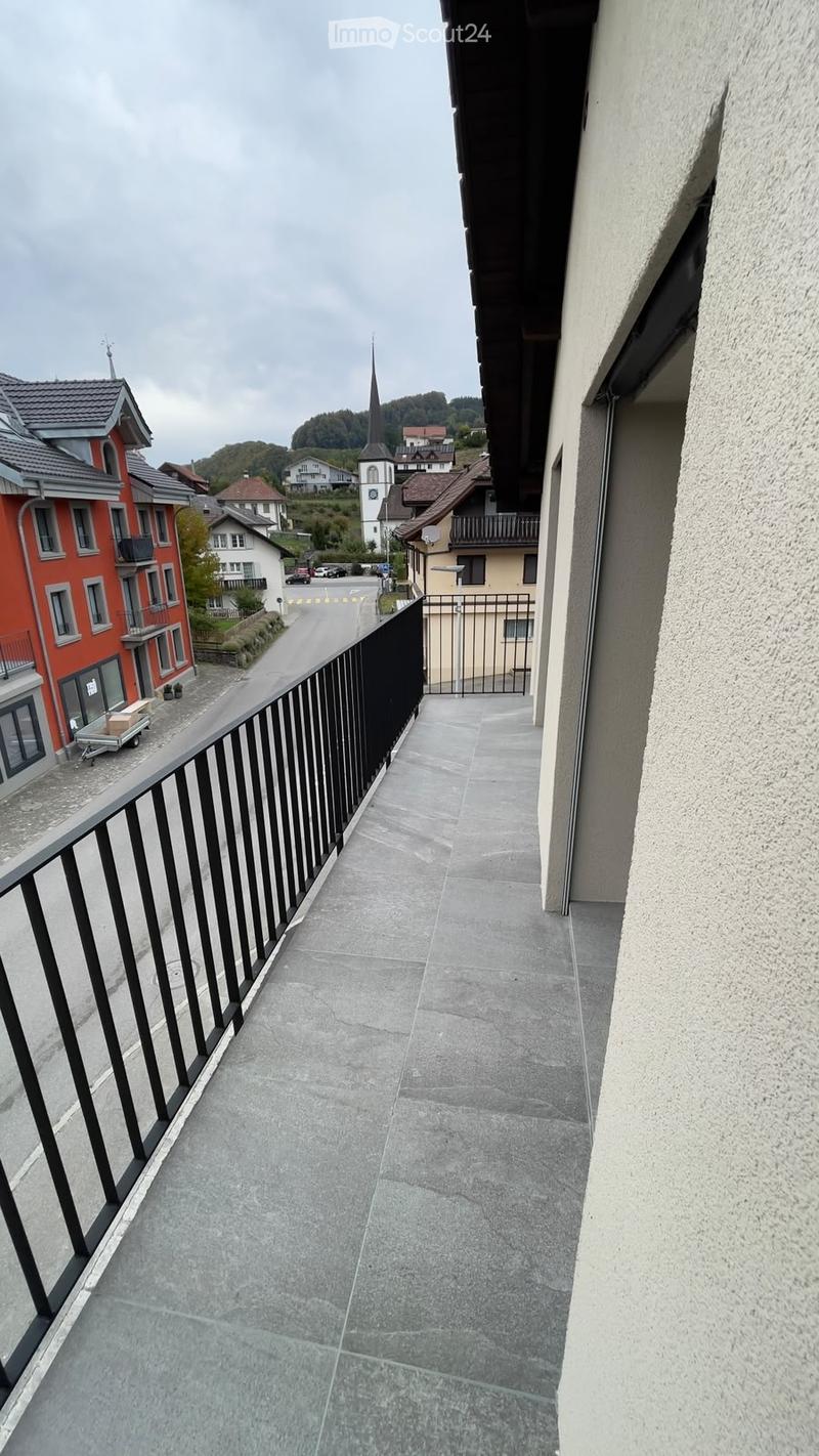 Balcony with grey tiled floor, black metal railing, overlooking a street with houses and a church in the background.