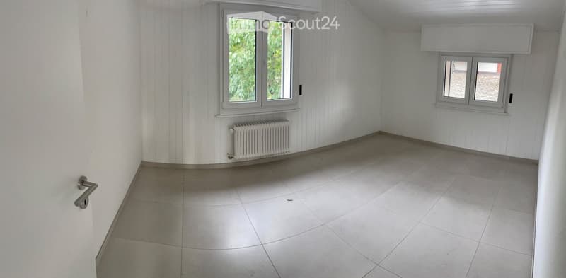 Empty room with white walls, tiled floor, two windows, radiator, and a door handle visible.