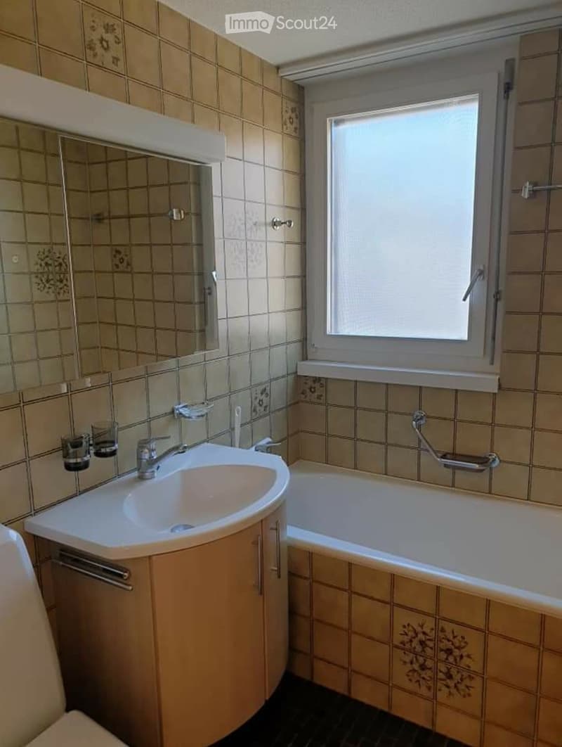Bathroom with a white sink, faucet, bathtub, mirror, and wooden cabinet. Beige tiled walls and a window with blinds.