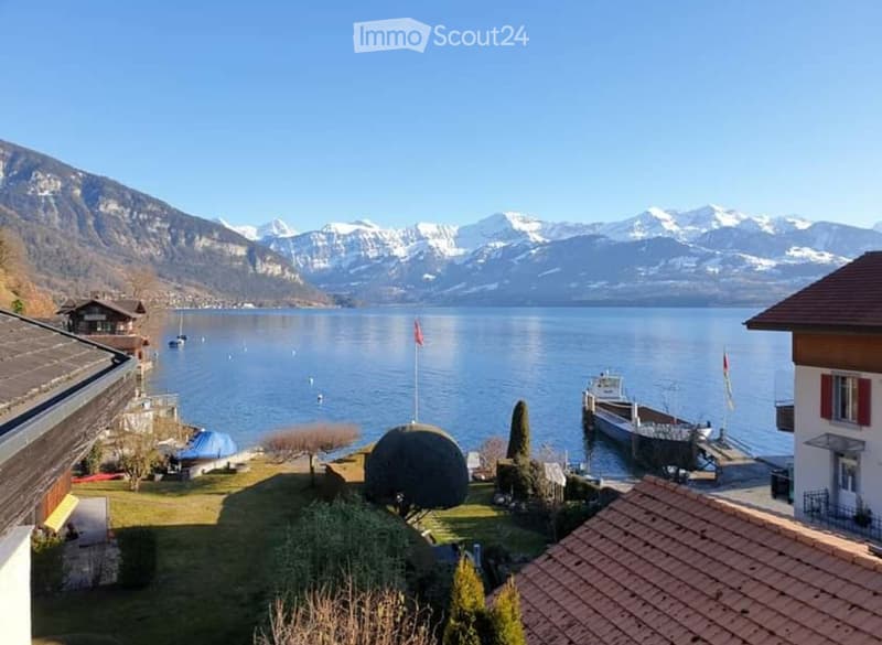 View of a lake with snow-capped mountains in the background, featuring a well-maintained garden area with trees, a flagpole, and a dock with boats.