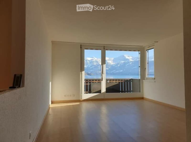 Empty room with wooden flooring, large windows offering a scenic mountain view, and a small balcony visible through the glass doors.