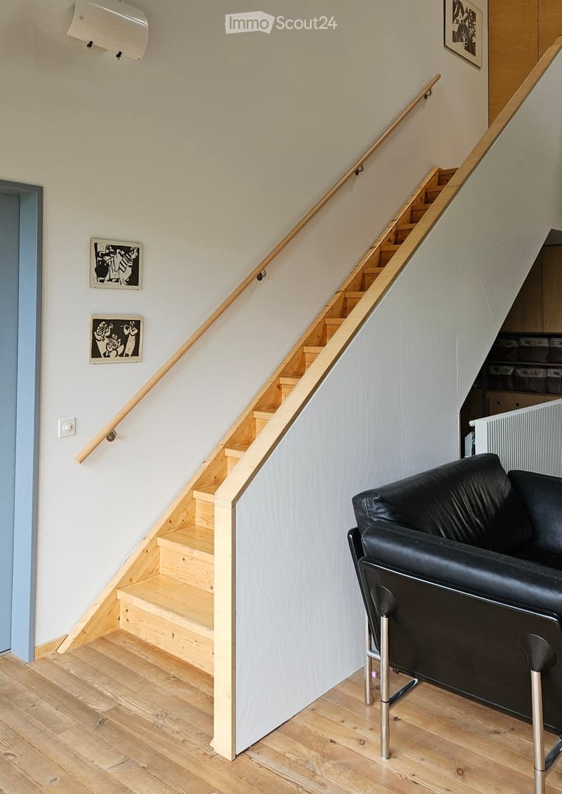A modern staircase with wooden steps and a metal railing is adjacent to a black leather sofa and a small metallic stool, with light wooden flooring and white walls.